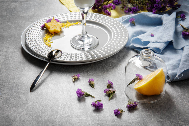 A bright set of flowers, plates and tropical fruits on a light gray table background. Blue and yellow fabric next to a plate with a tall glass, carambola and petals. Beautiful summer compositions.  - Photo, Image