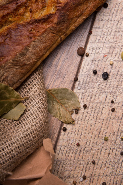A close-up picture of a crunchy, crusty, traditional french baguette on a rustic wooden background. Top view of a dry bay leaf and black round peppers with a dark brown baked pastry. - Foto, imagen