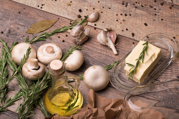 close-up picture of champignon mushrooms, spicy oil in a bottle, fresh rosemary and white garlic on a brown table background. Creamy butter in a glass box next to organic ingredients. - Photo, image