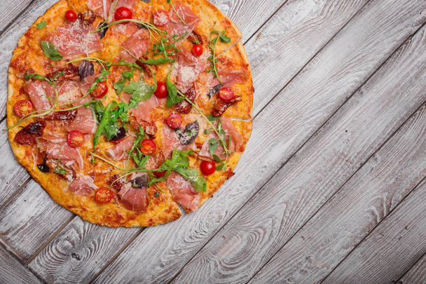 A perfectly round pizza with ham, spicy red pepper, fresh arugula, purple basil and hot grilled cheese on a gray wooden background, top view. Homemade mediterranean dinners for gourmets. Copy space. - Fotografie, Obrázek