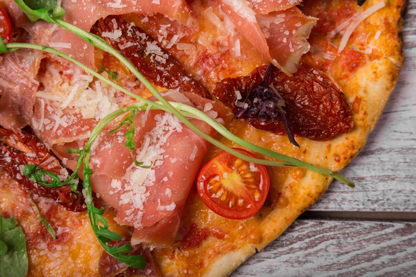 ein Makrobild frisch gekochter Pizza mit saftig rosa, salzigem Fleisch. Pizza mit geschnittenen Kirschtomaten, Salatblättern und gelbem Käse auf grauem Tischhintergrund. Koch- und Backkonzept. - Foto, Bild
