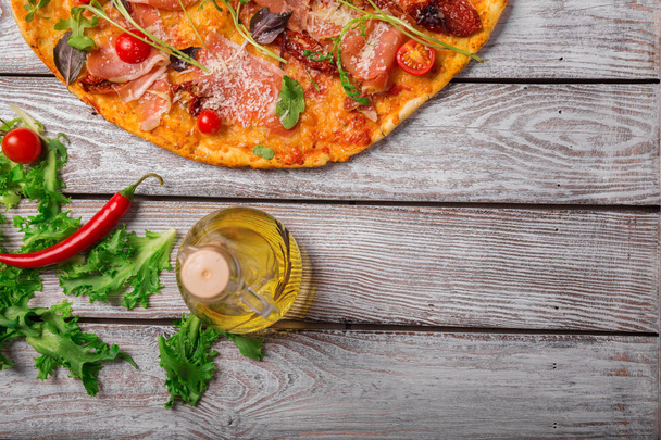 Italian cuisine. A view from above on a tasty pepperoni pizza with a bottle of olive oil and salad leaves. Delicious bright dish on a rustic wooden table background. Cooking concept. Copy space. - Fotoğraf, Görsel