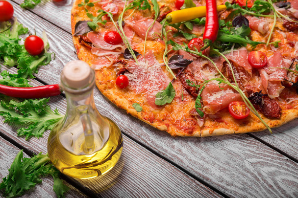 Un primer plano de una sabrosa pizza de margarita con verduras, una botella de aceite de oliva y hojas de ensalada. Colorido plato sobre un fondo rústico de mesa. Tomates cherry y chile. Concepto de cocina
. - Foto, Imagen