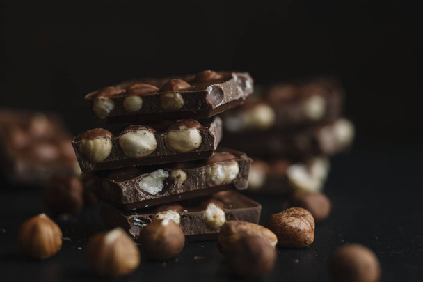 Chocolate bar pieces and nuts on dark slate background with copy space. Chocolate background. Top view. Toned - Zdjęcie, obraz