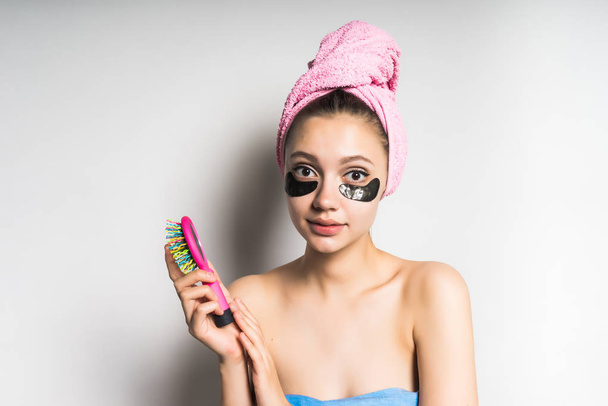 young beautiful girl with a pink towel on her head, silicone black patches under her eyes holding a comb in her hand - Foto, Imagen
