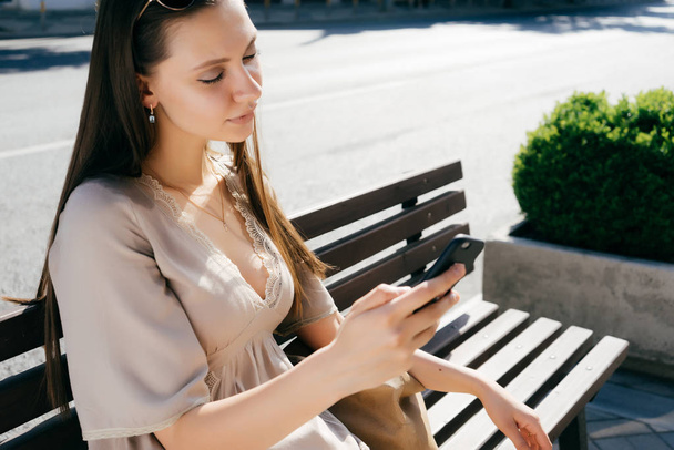 young beautiful long-haired girl in a beige dress sits on a wooden bench and looks in her smartphone - Foto, Imagem