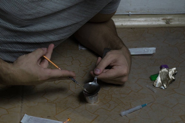 Um cara que é viciado em drogas se senta no chão e prepara uma dose em uma colher sobre uma vela
 - Foto, Imagem