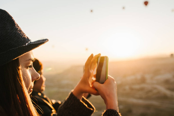 Zijaanzicht van charmante brunette meisje in hoed neemt afbeelding op smartphone tijdens vluchten van percelen van hete lucht ballonnen op warme zonnige dag. - Foto, afbeelding