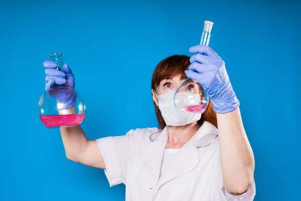 an adult female doctor in a protective medical mask and a white robe holds flasks with chemical solutions inside - Foto, Imagen