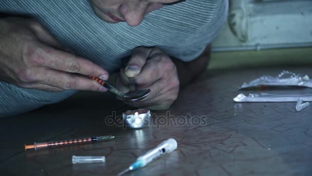 Een man die verslaafd aan de drugs is zit op de vloer en bereidt een dosis in een lepel op een kaars. - Video