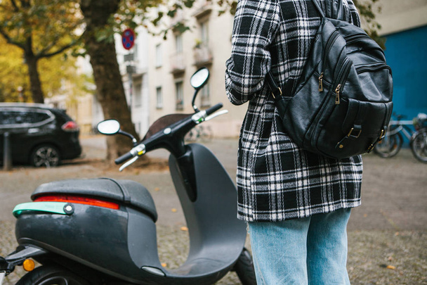Турист с рюкзаком собирается использовать электрический скутер через мобильное приложение в телефоне и дистанционно активировать его. Популярный автомобиль в Берлине и Париже
. - Фото, изображение
