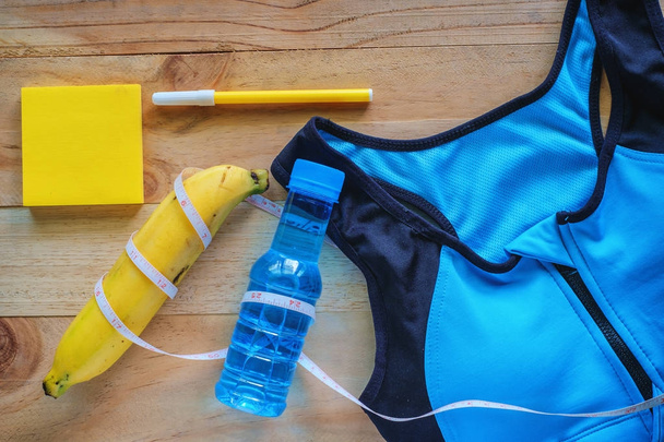 Top view of sport bra, fresh drinking water, banana, note pad, pen and measure tape on wooden table for exercise and fitness concept - Photo, Image