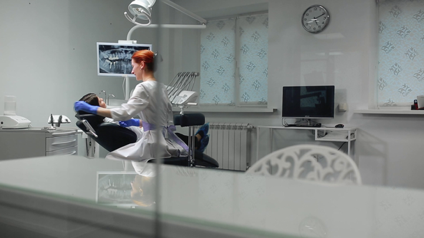 La doctora en guantes azules y una bata blanca se acerca a la silla del paciente y usando rayos X e instrumentos el dentista inspecciona los dientes en la cavidad oral. Chica paciente
. - Metraje, vídeo