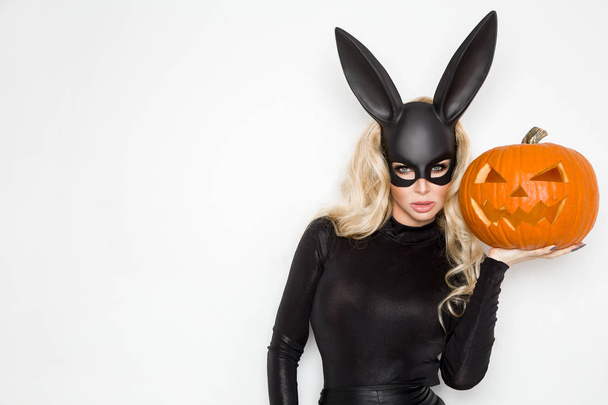 Beautiful young woman in halloween bunny costume, standing beside easter basket and holding pumpkin - Photo, Image