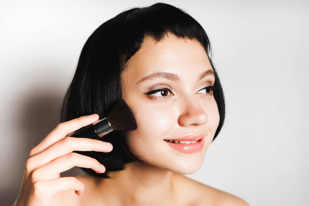 cute girl with short black hair puts powder on her cheeks with a makeup brush and looks away - Foto, Imagem