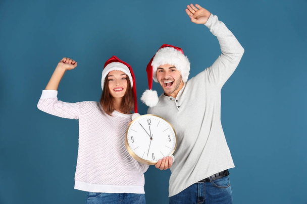 Young couple in Santa hats with clock on color background. Christmas countdown concept - Φωτογραφία, εικόνα