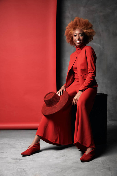 Amazing young black smiling model female with red hair wearing red wide pants, jacket, sweater and shoes sitting in studio with grey and red background and holding red hat - Fotó, kép