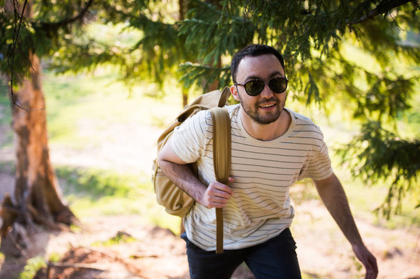 Komea tyylikäs nuori mies yllään valkoinen t-paita ja aurinkolasit reppu kädessään matkustaa metsässä. Poseeraus kameran edessä puun lähellä
 - Valokuva, kuva