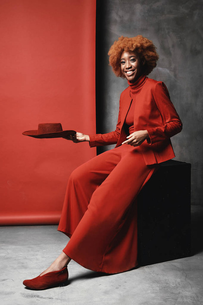 Dość młoda czarna kobieta uśmiechający się model z rude włosy, noszenie czerwony Szerokie spodnie, Kurtka, sweter oraz buty siedzi w studio z szarymi i czerwonymi tło i czerwony kapelusz gospodarstwa - Zdjęcie, obraz