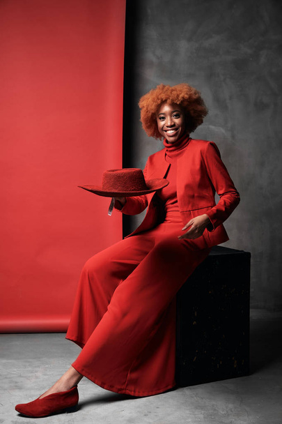 Pretty young black smiling model female with red hair wearing red wide pants, jacket, sweater and shoes sitting in studio with grey and red background with red hat - Φωτογραφία, εικόνα