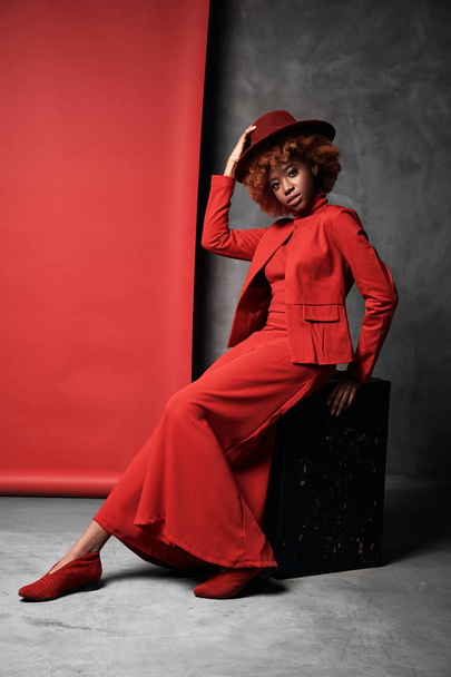 Pretty young black smiling model female with red hair wearing red wide pants, jacket, sweater and shoes sitting in studio with grey and red background with red hat - Fotó, kép