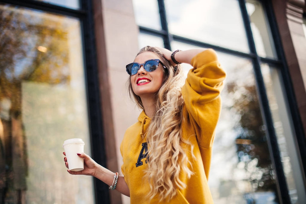 young beautiful blonde drinking coffee walking around the city.mustard sweetshot.,urban backpack , bright red lips Posing against the window of the boutique - Foto, Bild