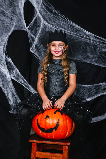Roztomilá holčička v halloween kostýmu sedí na dýně na pozadí s pavučina - Fotografie, Obrázek