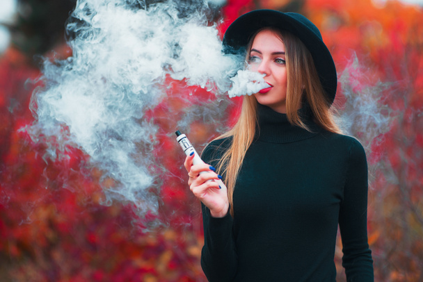 Vaping. Young beautiful woman in hat vaping outdoors in autumn - Photo, Image