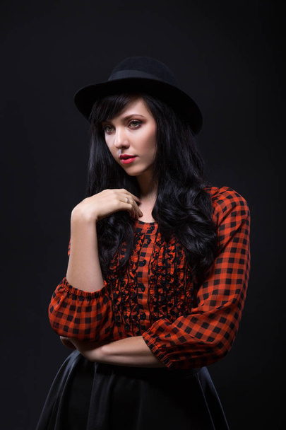 Συγκίνηση μελαχρινή γυναίκα μοντέλο ποζάρει με το μαύρο καπέλο με κόκκινο έντονο κραγιόν σε μαύρο φόντο. Τέχνη μόδα πορτρέτο. - Φωτογραφία, εικόνα