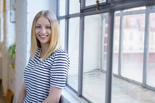 Young blonde smiling woman standing near window - Foto, afbeelding