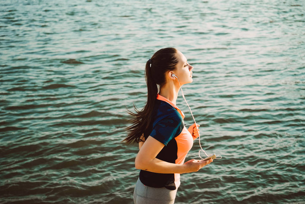 jovem bela menina esportiva correndo perto do rio ao pôr do sol, ouvindo música em fones de ouvido
 - Foto, Imagem