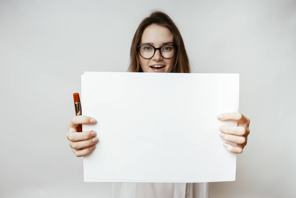 young beautiful girl in glasses smiles and holds in her hand a large sheet of white paper - Foto, imagen