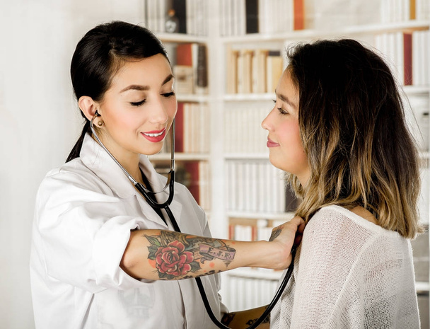 Bellissimo giovane medico tatuato utilizzando uno stestoscopio che controlla il torace del paziente, in background ufficio
 - Foto, immagini