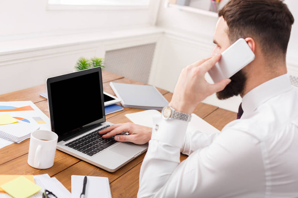 Серьезный бизнесмен разговаривает по мобильному телефону с ноутбуком в современном офисе
 - Фото, изображение