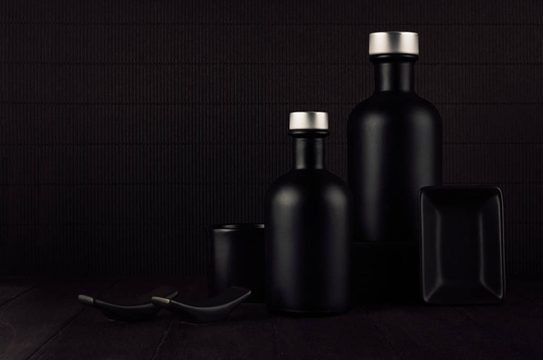 Dark black minimalist interior. Blank black cosmetics bottles on dark wood board, mock up.  - Фото, изображение