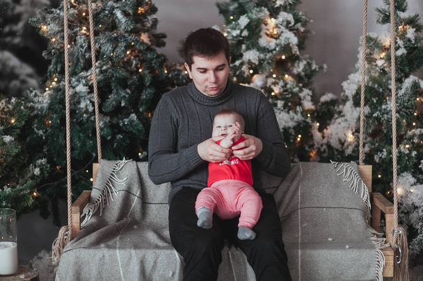 Father and 1 year old son playing in front of Christmas tree sitting on wooden swing in studio. - Foto, Imagem