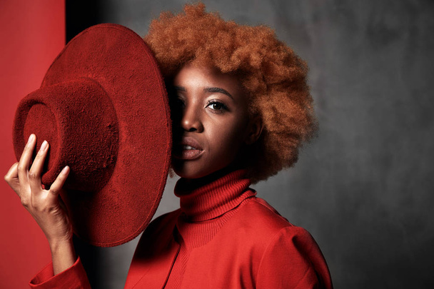 Portrait of beautiful sexy black female model wearing red sweater and jacket, holding red hat in her hand in studio with grey background - Фото, зображення