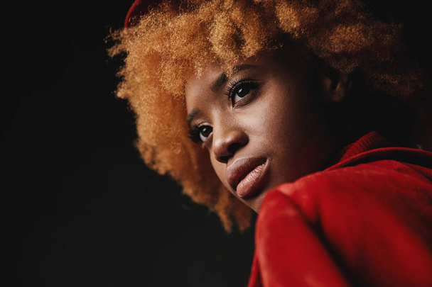 Closeup portrait of pretty sexy black female model wearing red sweater and jacket, with red curly hair in studio with grey background - Fotoğraf, Görsel