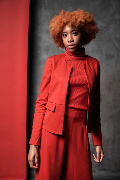 Young pretty black sexy woman with red curly short hair wearing red pants and jacket, posing in studio with red and grey background - 写真・画像