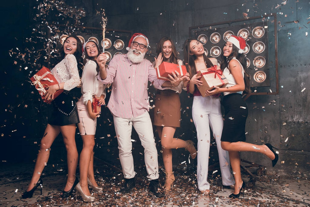 All they need is fun. Modern bearded man in Santa's hat carrying Christmas bengal lights with beautiful girls in club with gold confetti celebrate New Year and Christmas party 2018 - Photo, Image