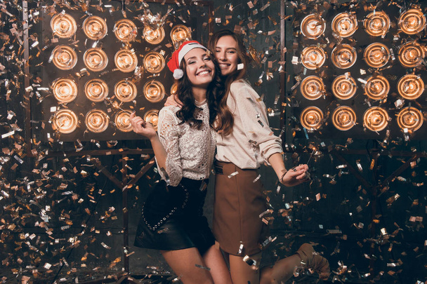 Twee gelukkige mooie meisjes in kerstmuts met Bengalen licht dansen en hebben een leuke in gouden en zilveren confetti. Kerstfeest met mijn beste vriend. New Year 2018 - Foto, afbeelding