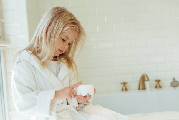 child in bathrobe applying face cream - Foto, Imagem