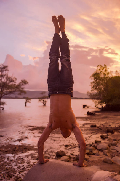Young Caucasian man is hand balancing on the rock at colorful sunset on background - Foto, imagen
