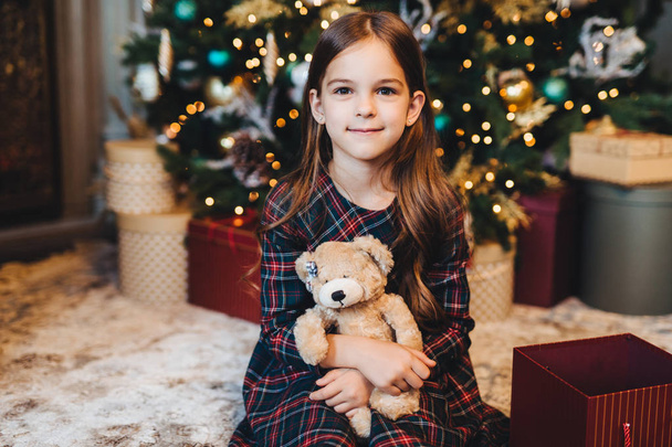 Small female child with appealing appearance, holds teddy bear, recieves present from parents, sits near Christmas tree. Adorable girl being glad to get gift on New Year. Christmas time. - Φωτογραφία, εικόνα