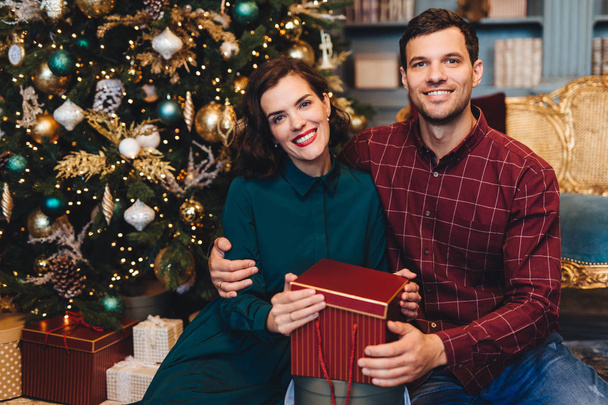 Šťastný nádherný pár v lásce, objali, sedí poblíž nastrojený stromeček nový rok, podržte přítomen, je rád, že slavit Vánoce společně. Manžel a manželka slaví Vánoce doma - Fotografie, Obrázek