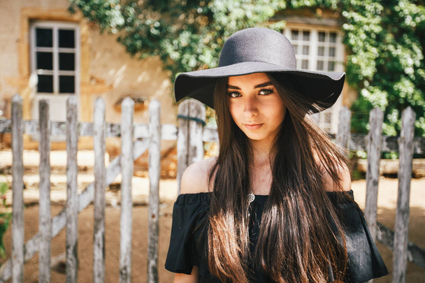 Menina sexy bonita morena com olhos castanhos em um vestido preto e um chapéu preto com grandes bordas contra o pano de fundo de uma cerca de madeira e uma velha casa em uma aldeia em uma Borgonha na frança
 - Foto, Imagem