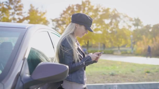 学生の女の子は sms のチャット、彼女のスマート フォンでメッセージを書く、晴れた秋の朝に彼女の黒い車の近くに立って - 映像、動画