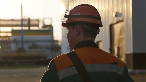 Slow motion backside view skilled worker in outfit walks along huge refinery territory under sunshine - Materiał filmowy, wideo