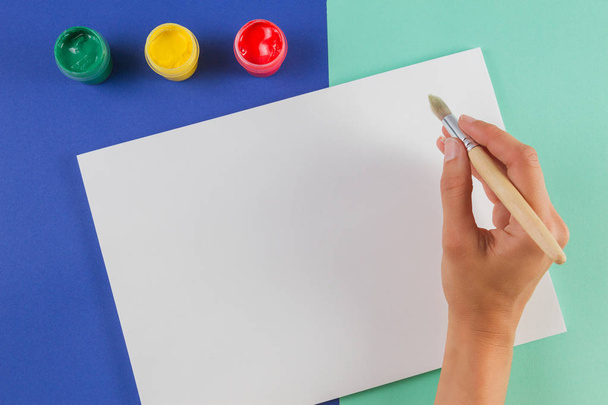 Γυναίκα χέρι με πινέλο ζωγραφικής, χρώματα watercolor και κενό φύλλο χαρτιού. - Φωτογραφία, εικόνα