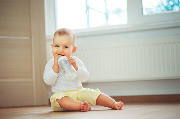 Little cute baby girl sitting in room on floor drinking water from bottle and smiling. Happy infant. Family people indoor Interior concepts. Childhood best time - Photo, Image